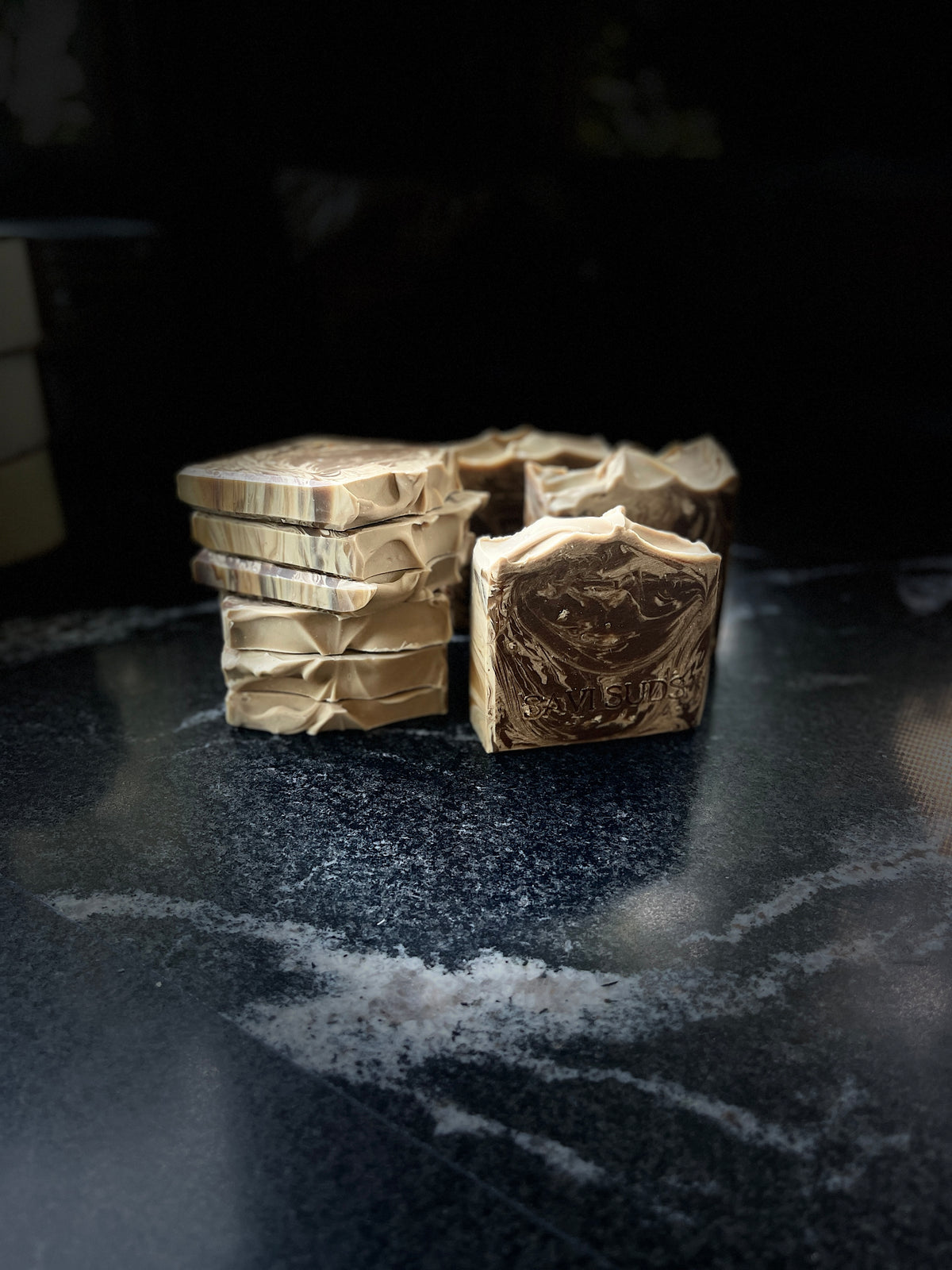 Several bars of handmade soap stacked on each other with one showing a dark brown swirl design, set on a dark marble surface.