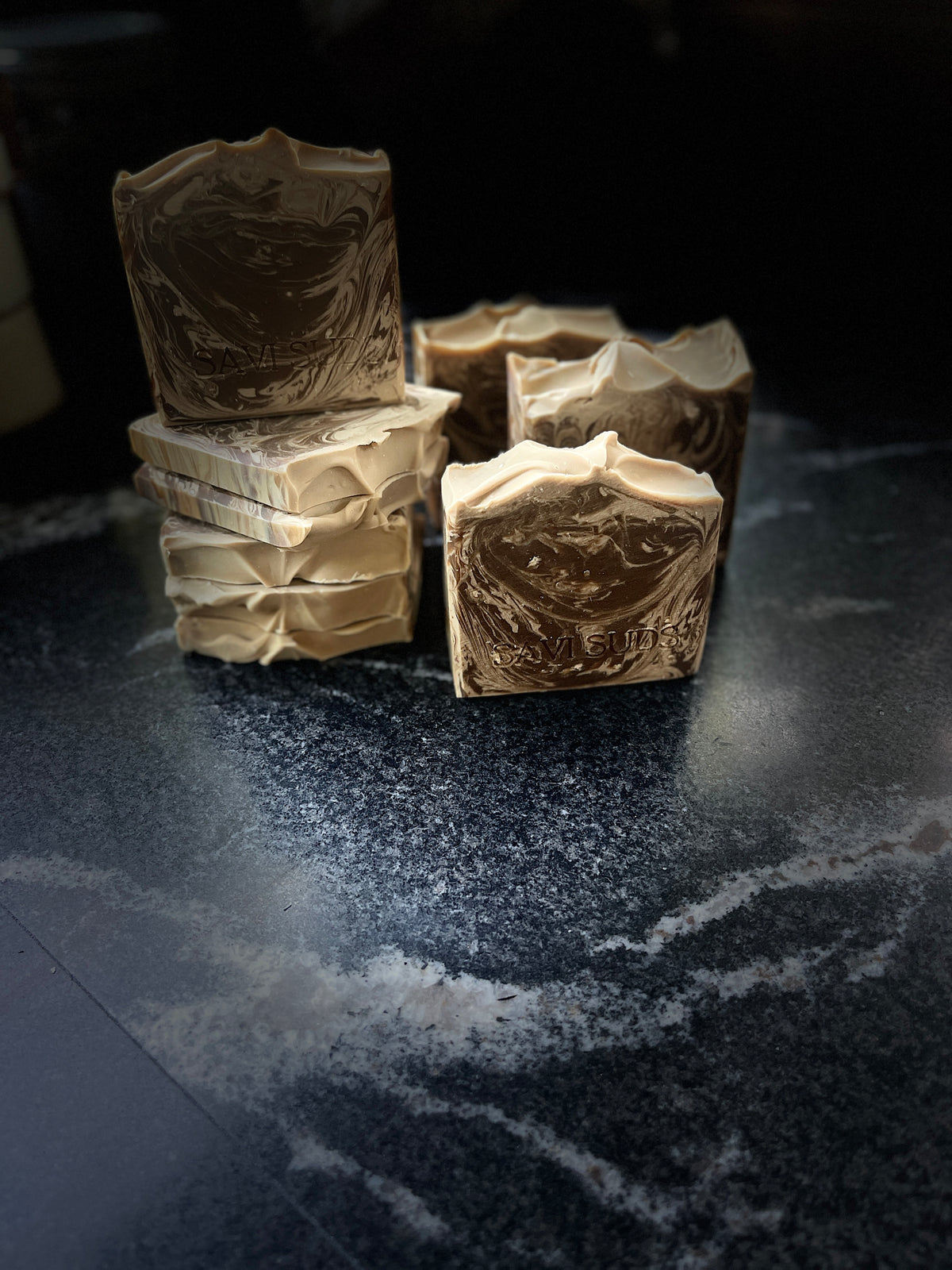 Several handmade square bars of soap with brown and cream swirls, arranged on a dark marble-like surface against a dark background.
