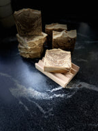 Several handmade soap bars with a brown and beige swirls are arranged on a dark stone surface, with one bar in the foreground resting on a small wooden stand. The soap has the brand name 'SAVI SUDS' stamped on the front