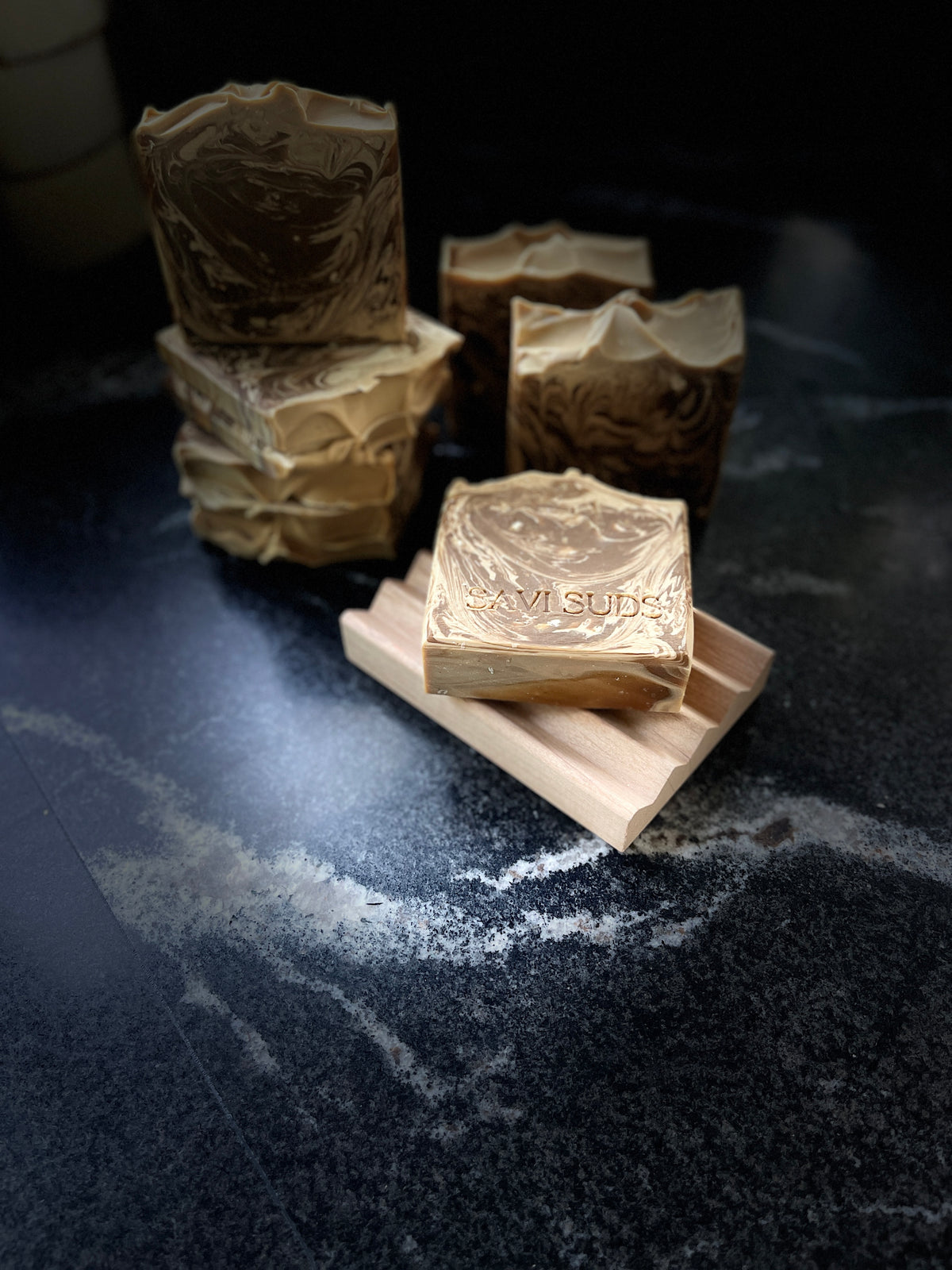 Several handmade soap bars with a brown and beige swirls are arranged on a dark stone surface, with one bar in the foreground resting on a small wooden stand. The soap has the brand name 'SAVI SUDS' stamped on the front