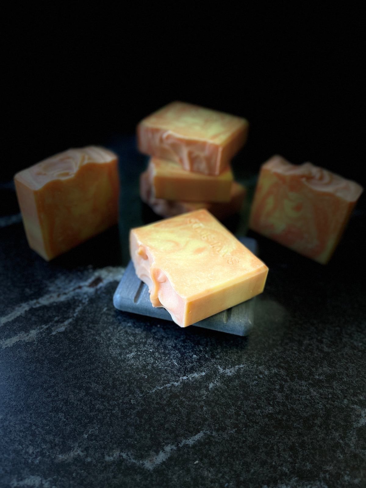 Several square bars of marbled orange and cream soap arranged on a dark textured surface against a black background.