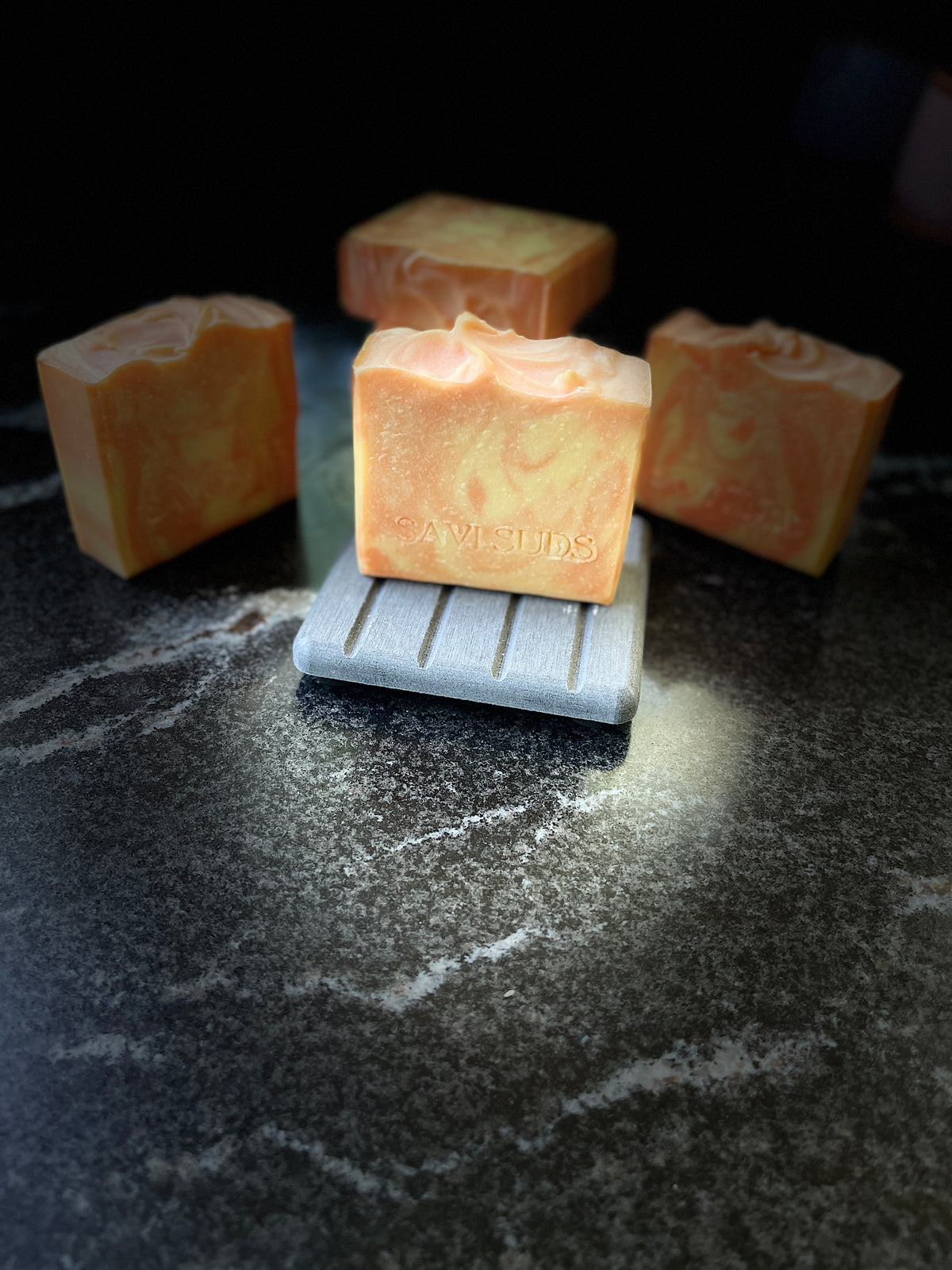 Four orange and cream marbled artisanal soap bars arranged on a dark stone surface, with one bar centered on a small gray slotted dish. The center bar has the brand name 'SAVI SUDS' stamped on it.