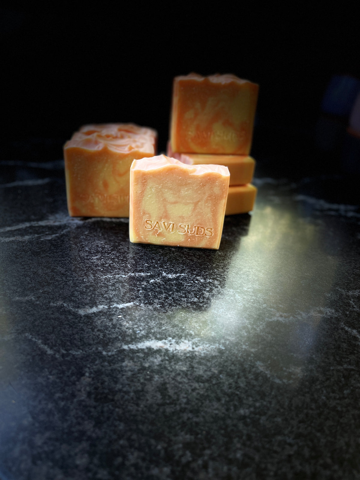 Four orange-colored handmade soap bars with 'SAVI SUDS' stamped on the front, arranged on a dark, reflective marble-like surface against a black background.