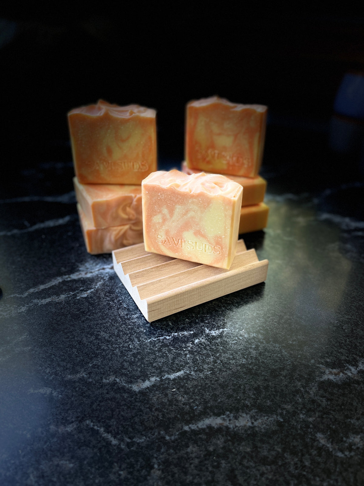 Four handmade marbled soap bars in orange and cream tones, with one bar displayed on a small wooden stand in the foreground against a dark marble surface and black background.