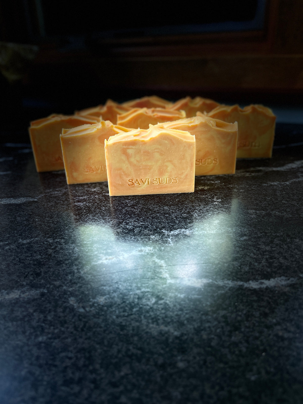 Multiple orange and cream colored handmade soap bars with 'SAVI SUDS' stamped on the front, arranged on a dark, reflective marble-like surface against a black background.