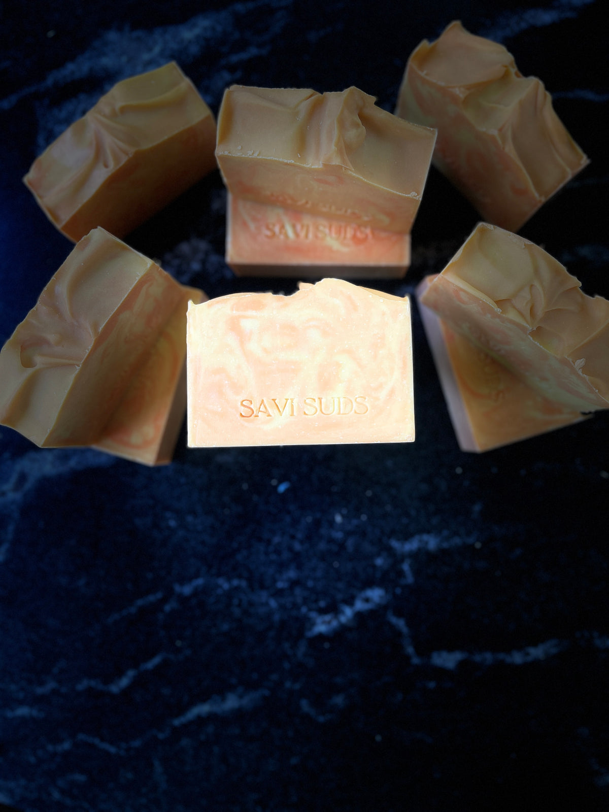 Multiple orange and cream colored handmade soap bars with 'SAVI SUDS' stamped on the front, arranged on a dark, reflective marble-like surface against a black background.