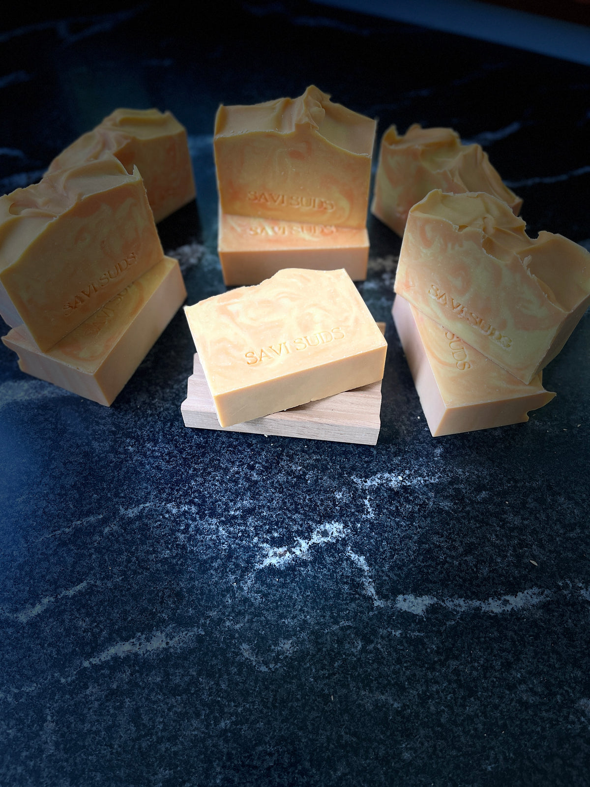 Multiple orange and cream colored handmade soap bars with 'SAVI SUDS' stamped on the front, arranged on a dark, reflective marble-like surface against a black background.