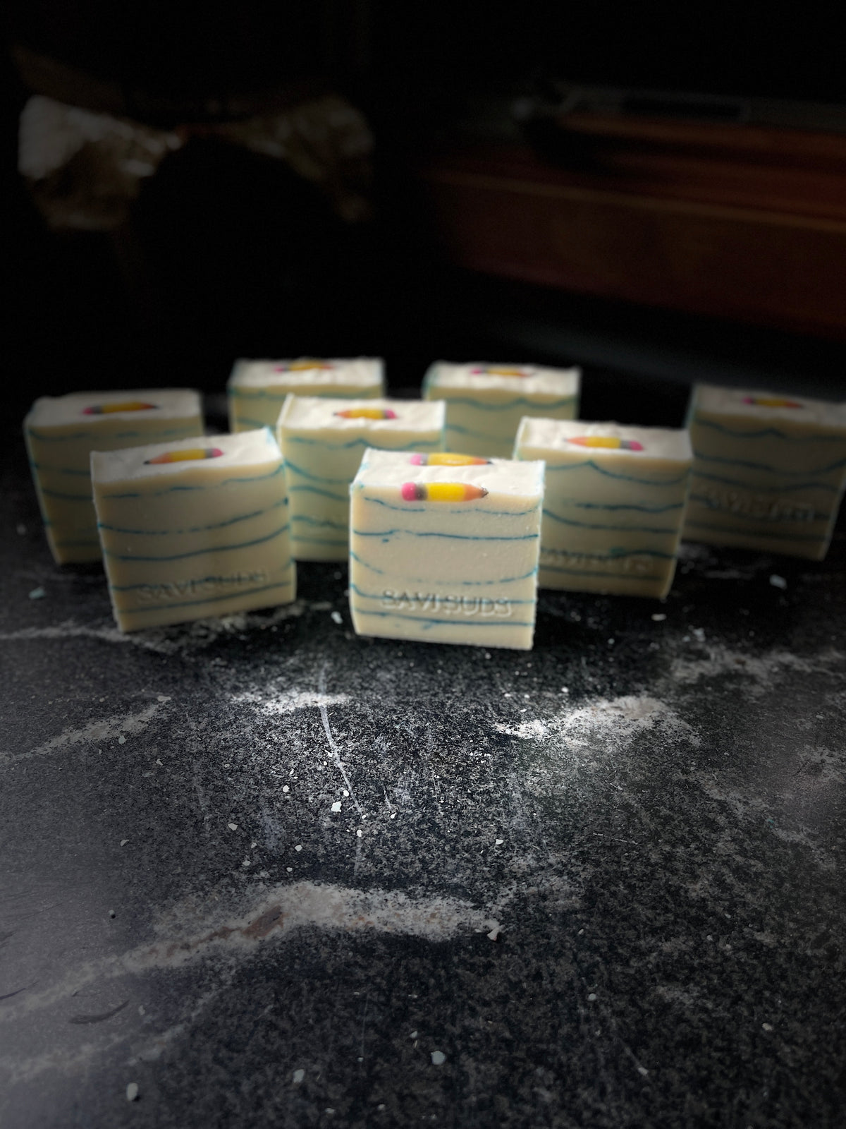Several square bars of handmade soap with white layers, blue mica line, and a pencil embed on top. Soap is arranged on a dark marble surface