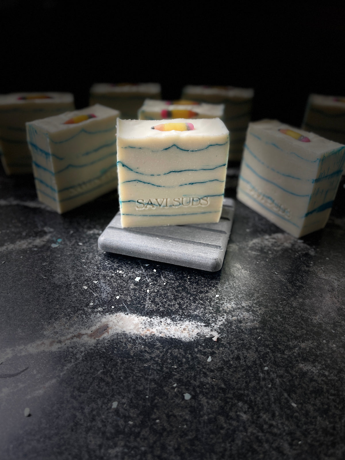 A square bar of handmade soap with white layers, blue wavy lines, and a small pencil embed on top, displayed on a gray stone coaster. The soap bar has 'Savi Suds' printed on the front, with other soap bars blurred in the background on a dark textured surface.