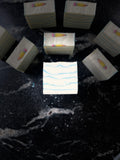 A set of white square soap bars with light blue wavy line patterns and small yellow-and-pink pencil embeds on top, arranged on a dark black marble surface. One bar in the center has faint text reading 'SAVI SUDS'.