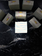 A set of white square soap bars with light blue wavy line patterns and small yellow-and-pink pencil embeds on top, arranged on a dark black marble surface. One bar in the center has faint text reading 'SAVI SUDS'.