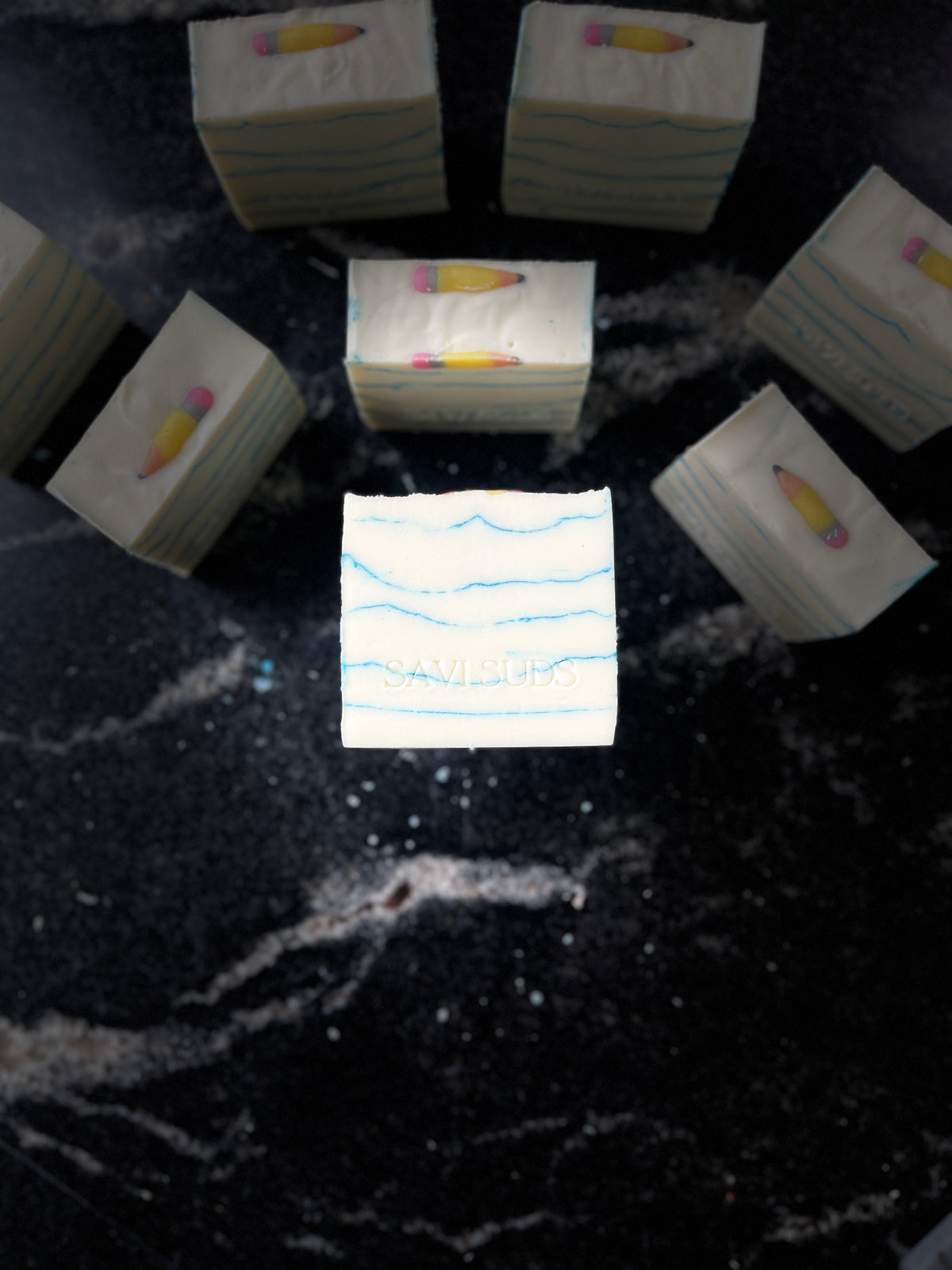 A set of white square soap bars with light blue wavy line patterns and small yellow-and-pink pencil embeds on top, arranged on a dark black marble surface. One bar in the center has faint text reading 'SAVI SUDS'.