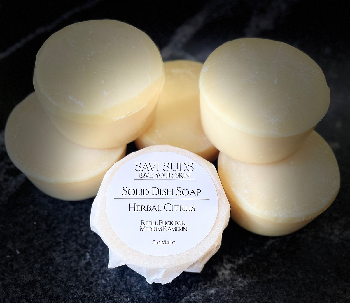 Multiple round bars of Savi Suds Solid Dish Soap in Herbal Citrus scent, with one bar in the foreground wrapped in paper showing the white label, set on a dark textured surface.
