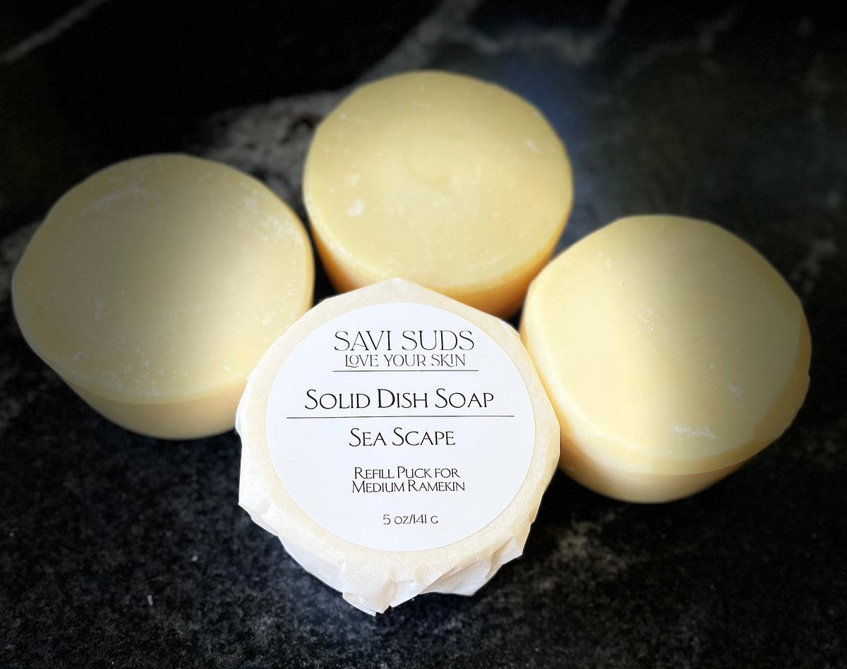 Four round bars of Savi Suds Solid Dish Soap in Sea Scape scent, with one bar in the foreground wrapped in paper showing the white label, set on a dark textured surface.