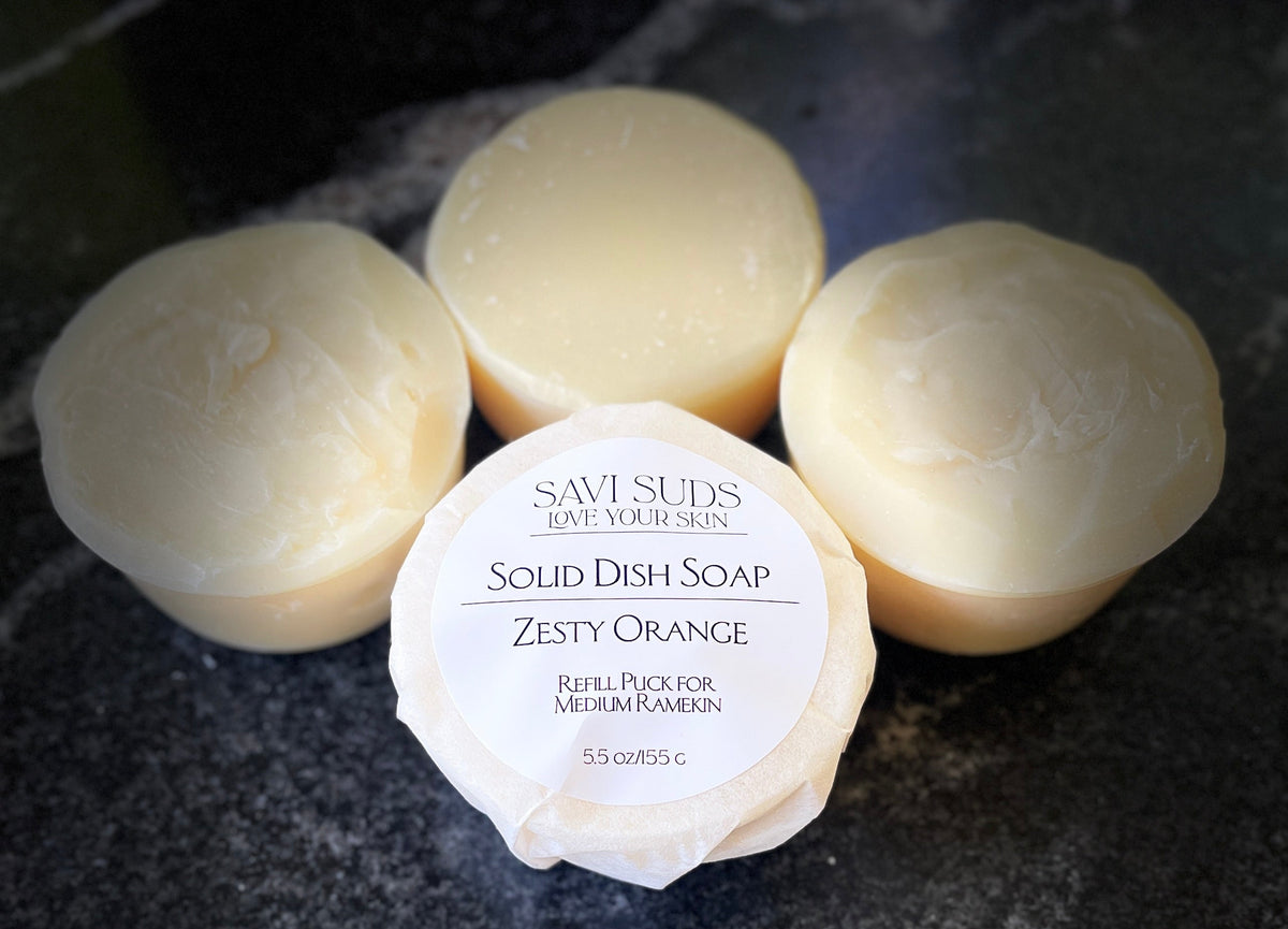 Four round bars of Savi Suds Solid Dish Soap in Zesty Orange scent, with one bar in the foreground wrapped in paper showing the white label, set on a dark textured surface.