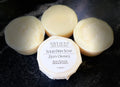 Four round bars of Savi Suds Solid Dish Soap in Zesty Orange scent, with one bar in the foreground wrapped in paper showing the white label, set on a dark textured surface.