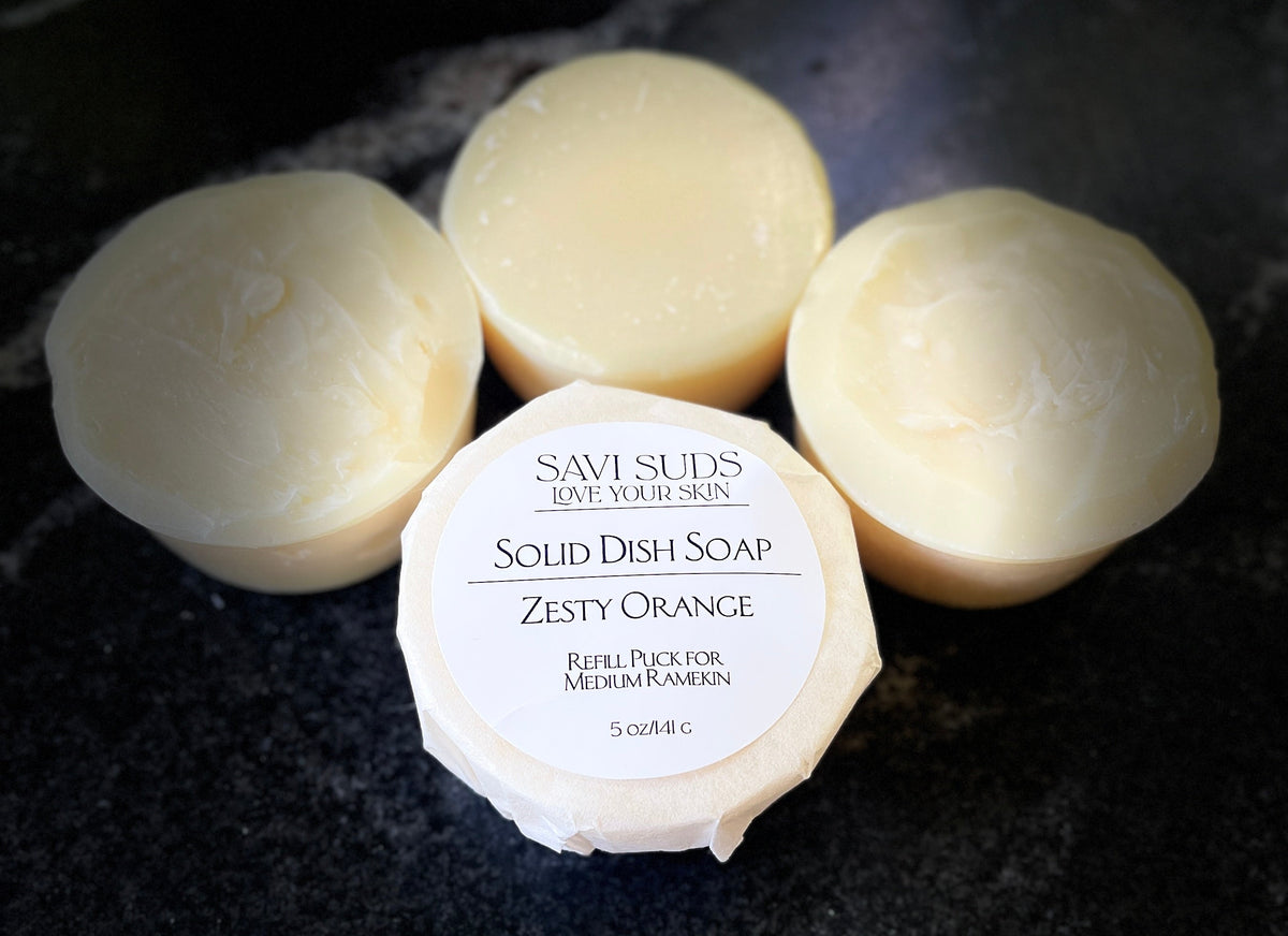Four round bars of Savi Suds Solid Dish Soap in Zesty Orange scent, with one bar in the foreground wrapped in paper showing the white label, set on a dark textured surface.