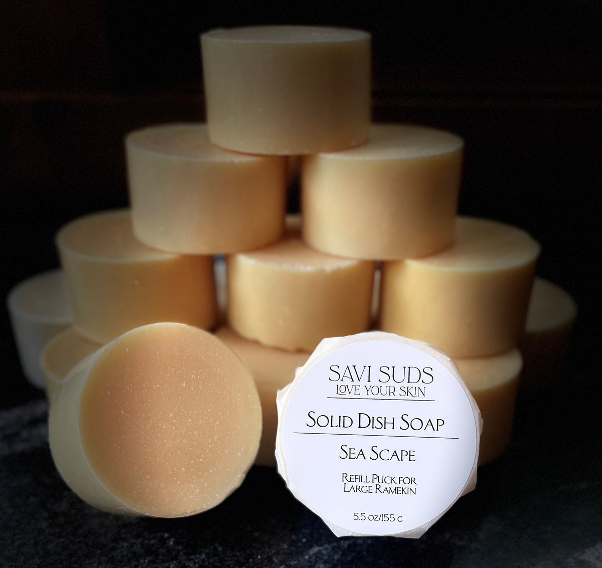 A stack of solid dish soap refill bars with one in front wrapped in bleach free coffee filter that reads 'SAVI SUDS' and 'SOLID DISH SOAP SEA SCAPE', set on a dark textured countertop.