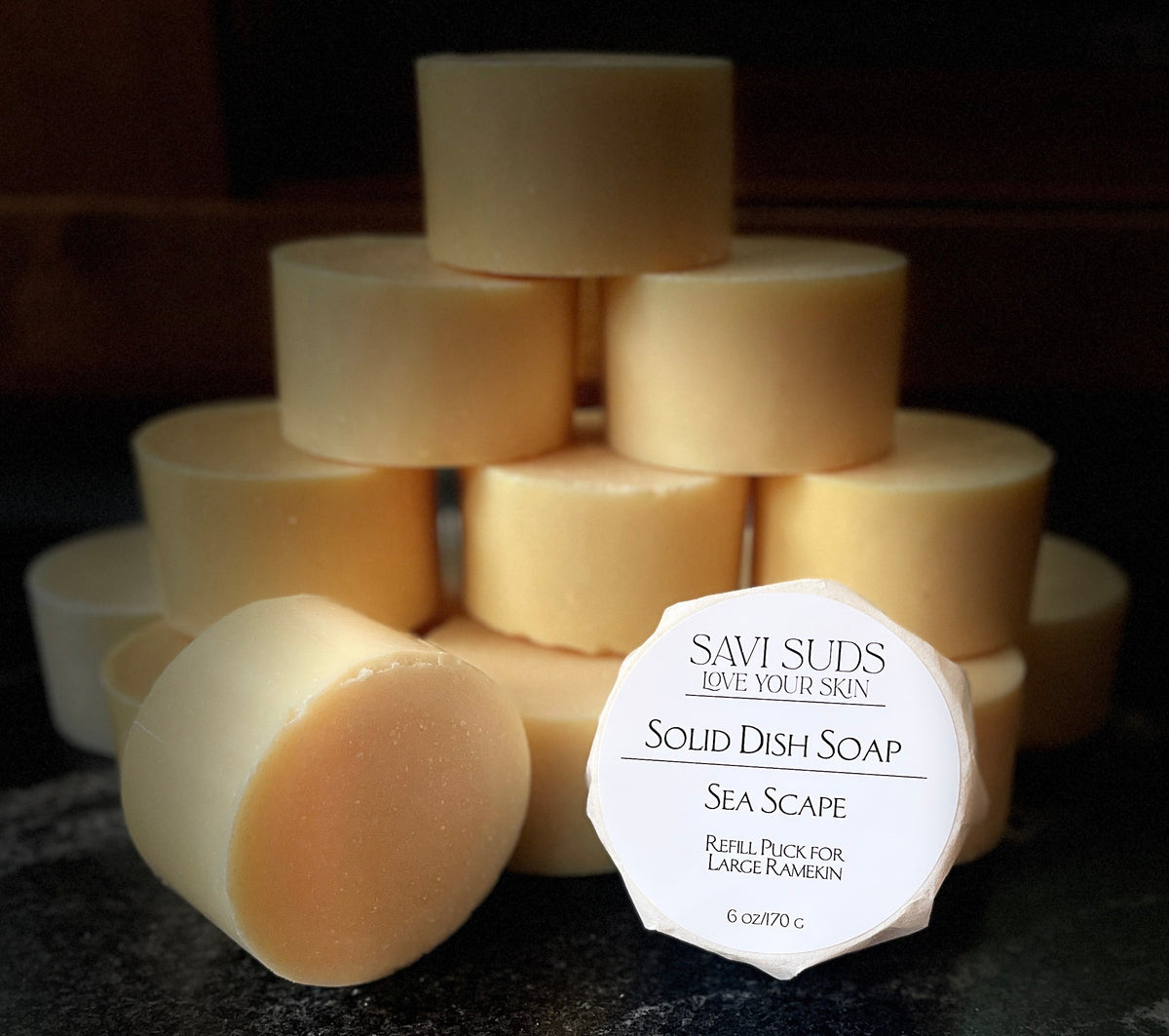 A stack of solid dish soap refill bars with one in front wrapped in bleach free coffee filter that reads 'SAVI SUDS' and 'SOLID DISH SEA SCAPE', set on a dark textured countertop.