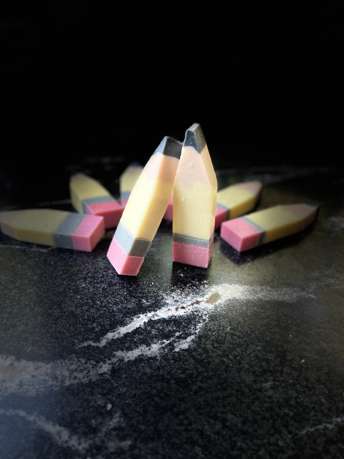 Three novelty pencil-shaped soaps standing upright on a dark textured surface, with several more lying in the background. The soaps are designed to look like pencils, featuring pale yellow bodies, gray and black sharpened tips, and pink erasers.