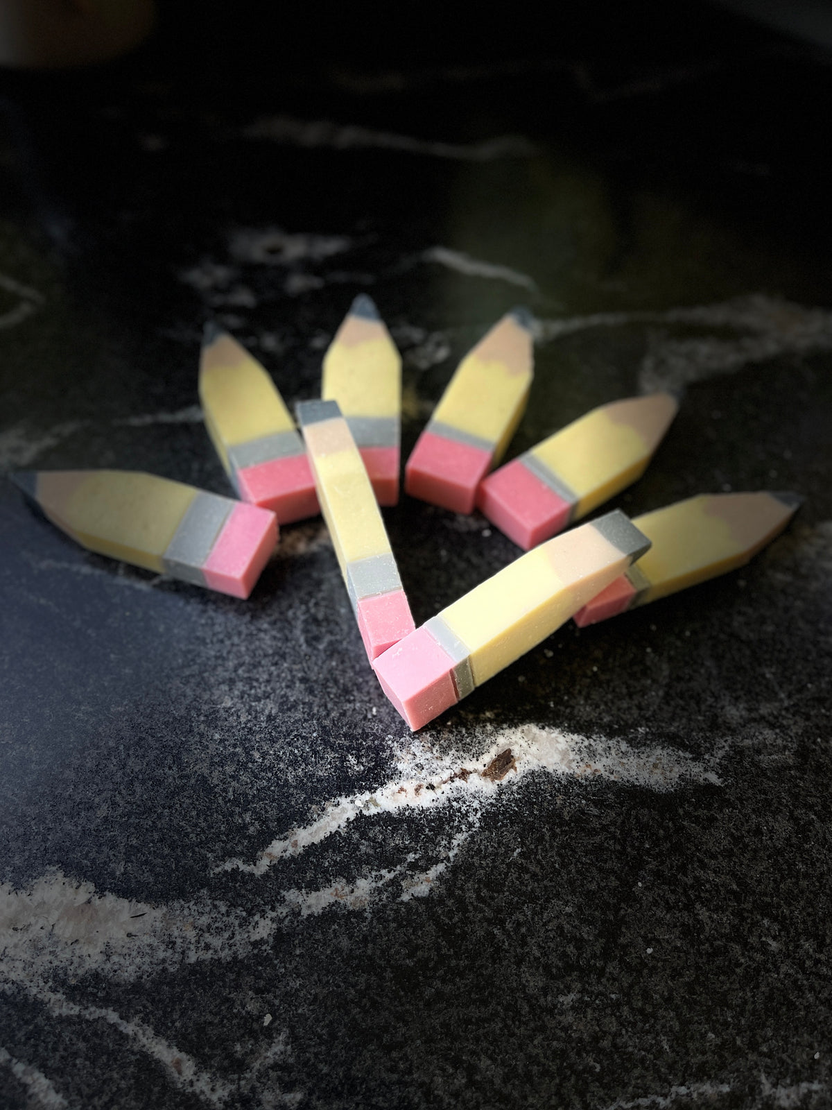 Five miniature yellow pencil-shaped soaps arranged in a loose circle on a dark, textured stone surface.