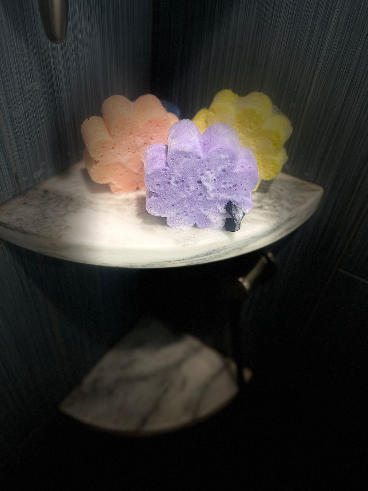 Three flower-shaped bath sponges in pastel orange, purple, and yellow, displayed on a curved white marble shelf inside a shower