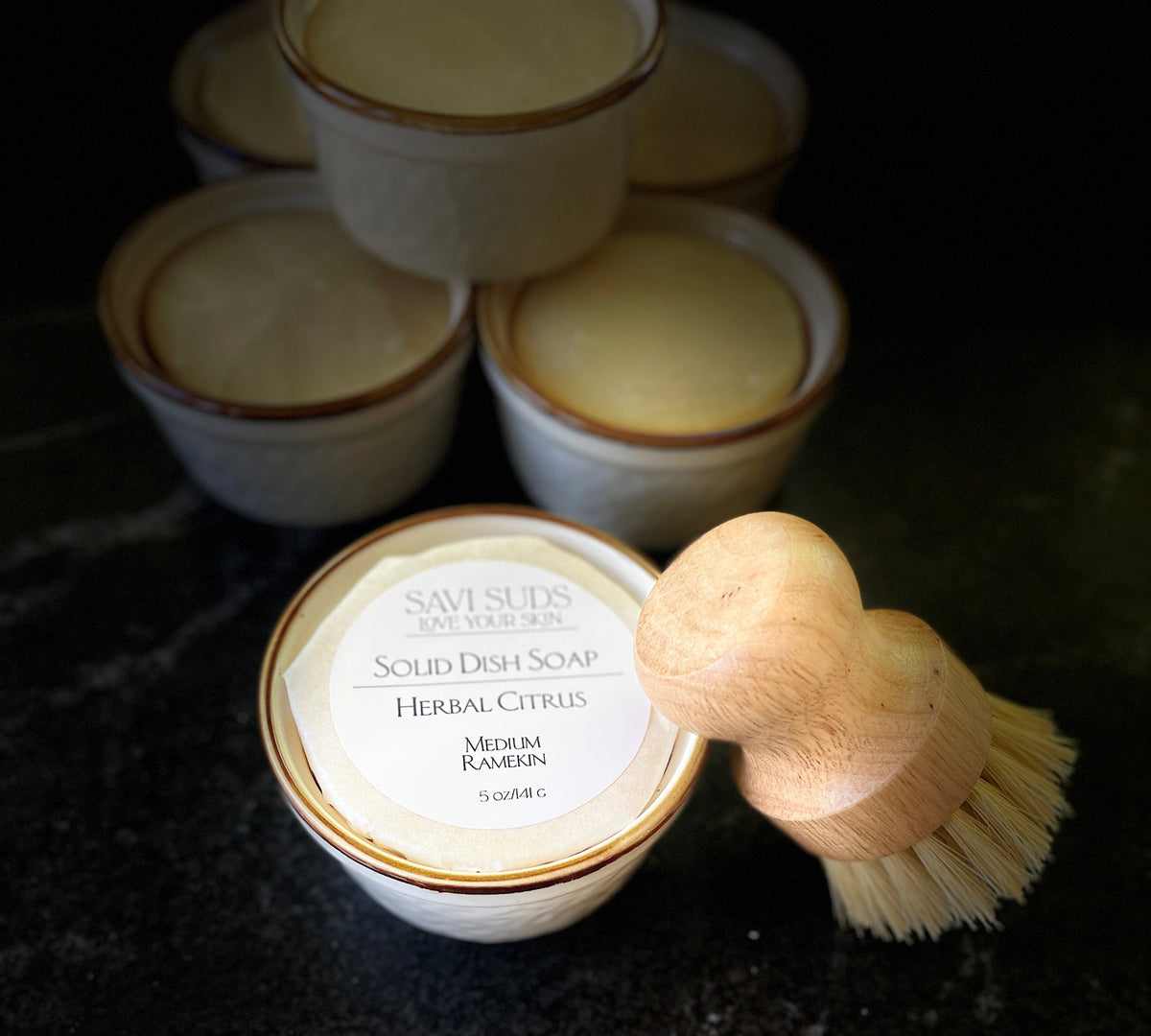 A set of small white ceramic dish soap containers with brown trim on a dark marble countertop, with a wooden-handled brush resting on the front container labeled 'Savi Suds Solid Dish Soap Herbal Citrus'.