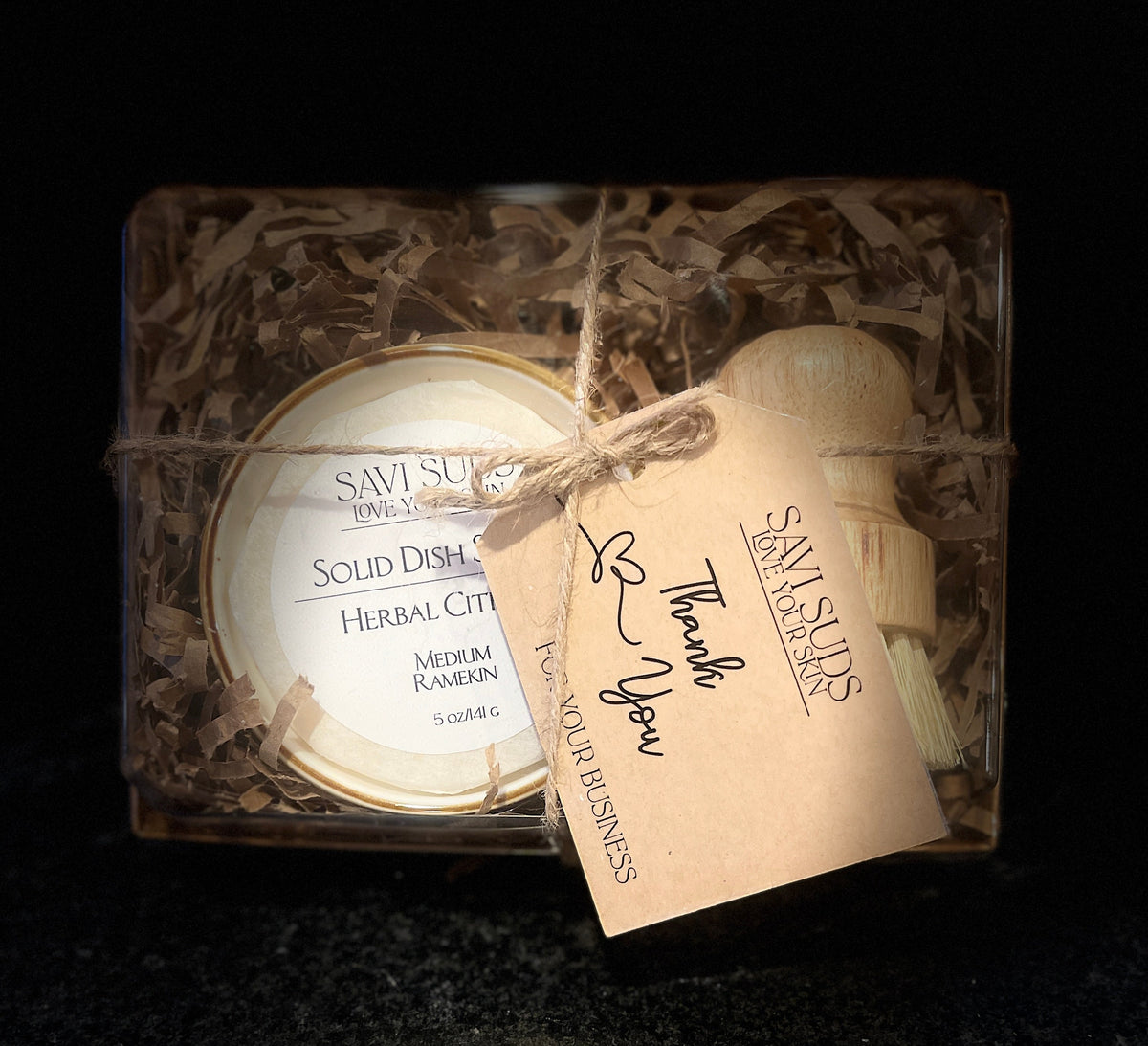 A boxed dishwashing brush set from Savi Suds, featuring a round solid dish soap in a ramekin, a small bamboo dish brush, and a kraft paper thank you tag, all wrapped in twine inside a clear plastic box with shredded paper filler.