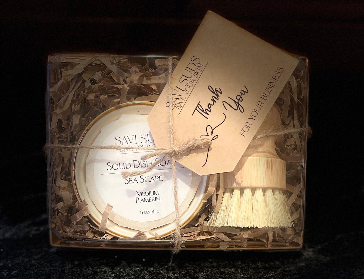 A boxed dishwashing brush set from Savi Suds, featuring a round solid dish soap in a ramekin, a small bamboo dish brush, and a kraft paper thank you tag, all wrapped in twine inside a clear plastic box with shredded paper filler.