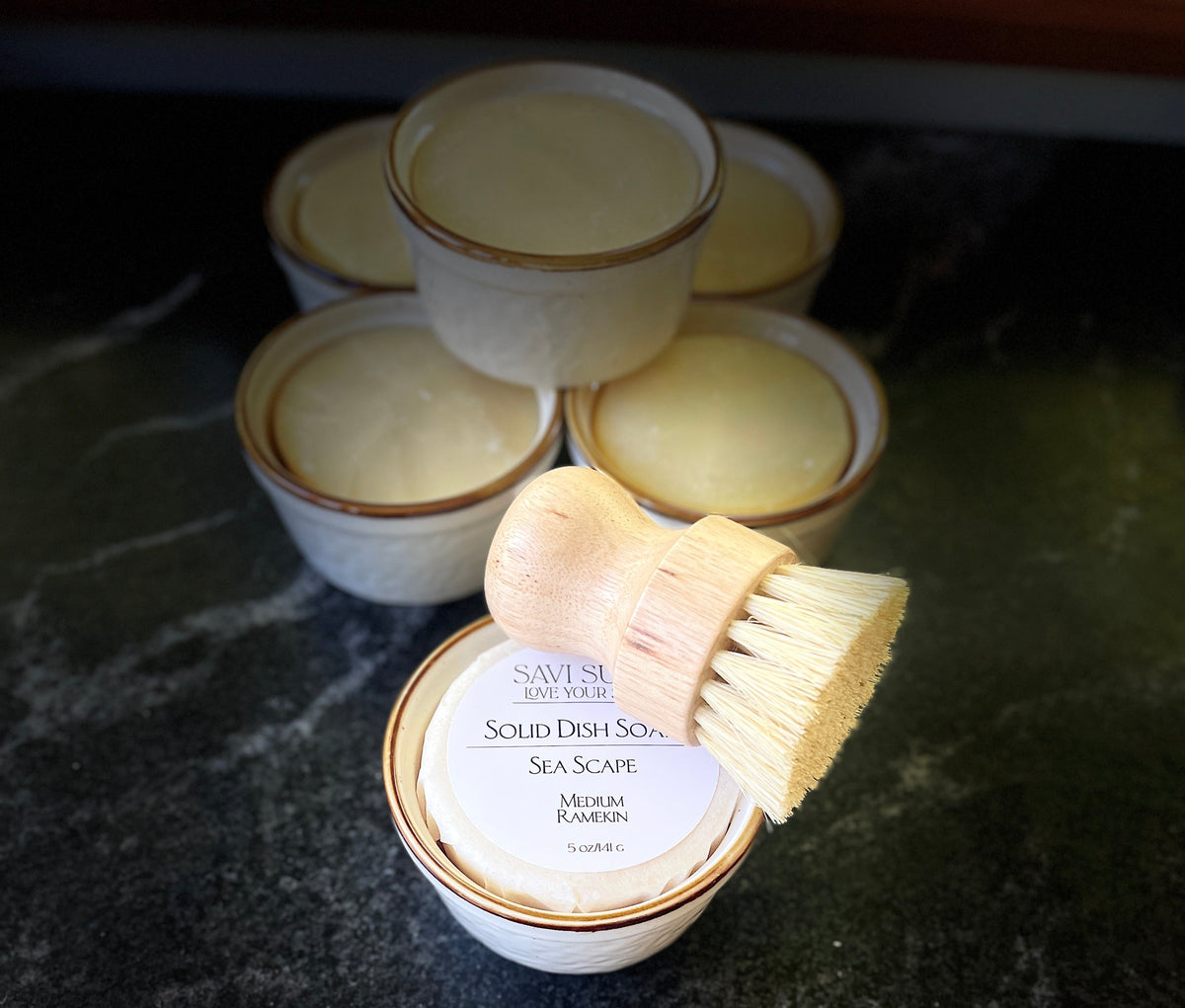 A set of small white ceramic dish soap containers with brown trim on a dark marble countertop, with a wooden-handled brush resting on the front container labeled 'Savi Suds Solid Dish Soap Sea Scape'.