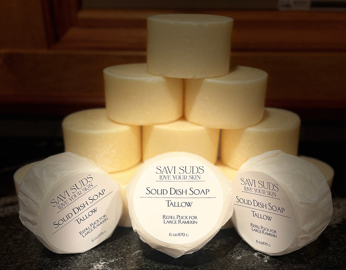 Three wrapped bars of Savi Suds Tallow Solid Dish Soap arranged on a dark countertop, with multiple bars stacked behind them in a pyramid.