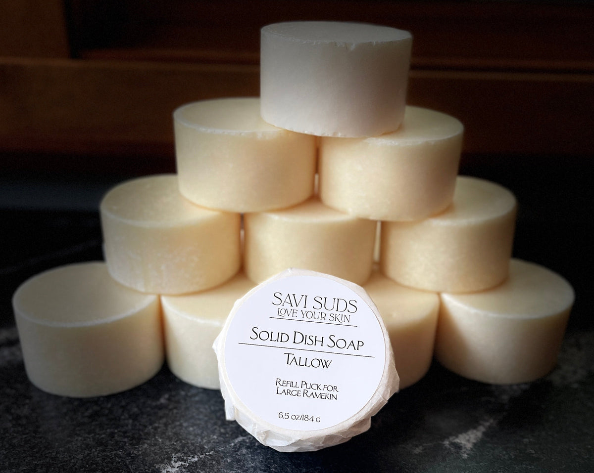 A stack of round, pale off-white solid dish soap bars arranged in a pyramid on a dark countertop, with one soap bar in the front center wrapped in a bleach free coffee filter and labeled 'SAVI SUDS Solid Dish Soap Tallow'.
