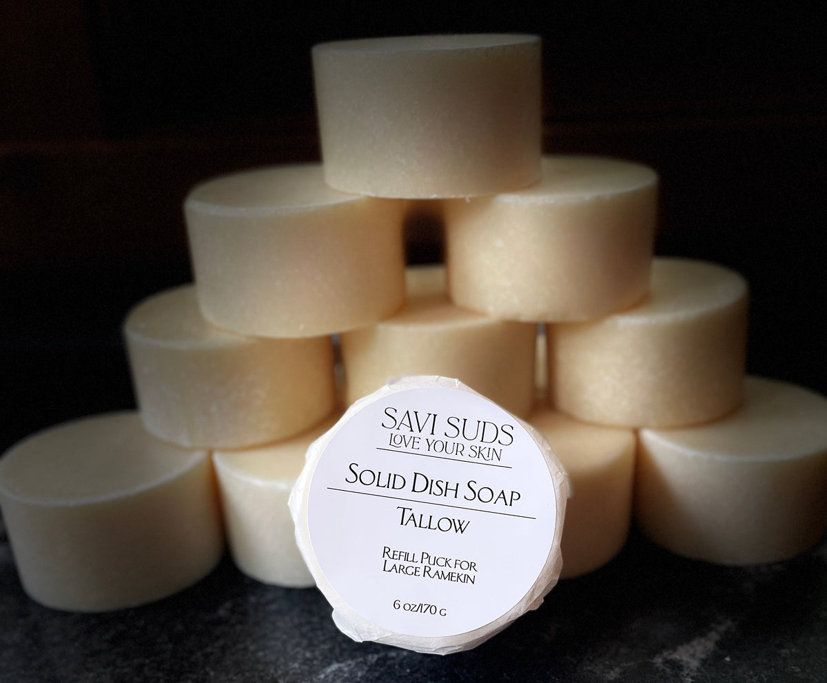 A stack of cream-colored solid dish soap bars arranged in a pyramid, with one bar in the front displaying its white paper label that reads 'SAVI SUDS SOLID DISH SOAP TALLOW'. The soaps are photographed on a dark, textured countertop against a dark background.