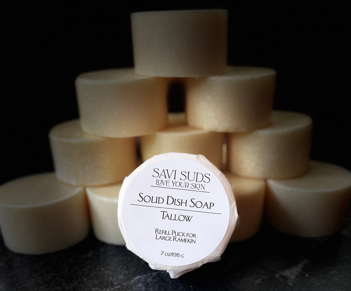 A round bar of Savi Suds Solid Dish Soap in Tallow, wrapped in bleach free coffee filter with the brand name clearly visible, set in front of a pyramid stack of similar soap bars on a dark, textured surface.