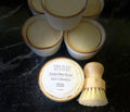 A set of small white ceramic dish soap containers with brown trim on a dark marble countertop, with a wooden-handled brush resting on the front container labeled 'Savi Suds Solid Dish Soap Zesty Orange'.