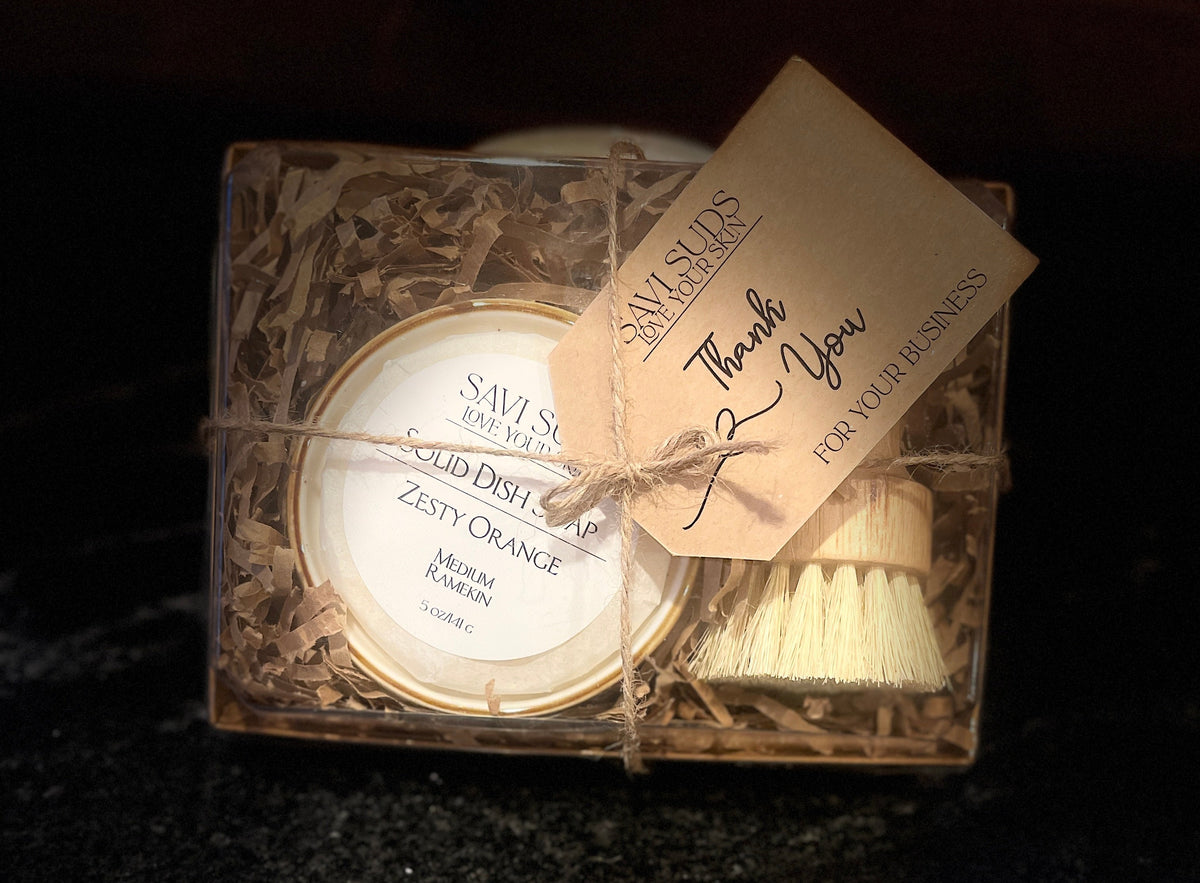 A boxed dishwashing brush set from Savi Suds, featuring a round solid dish soap in a ramekin, a small bamboo dish brush, and a kraft paper thank you tag, all wrapped in twine inside a clear plastic box with shredded paper filler.