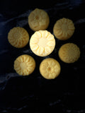 Seven pale yellow flower-shaped soap bars arranged on a dark marble-like surface, with one central soap bar highlighted by brighter lighting.