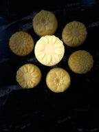 Seven pale yellow flower-shaped soap bars arranged on a dark marble-like surface, with one central soap bar highlighted by brighter lighting.
