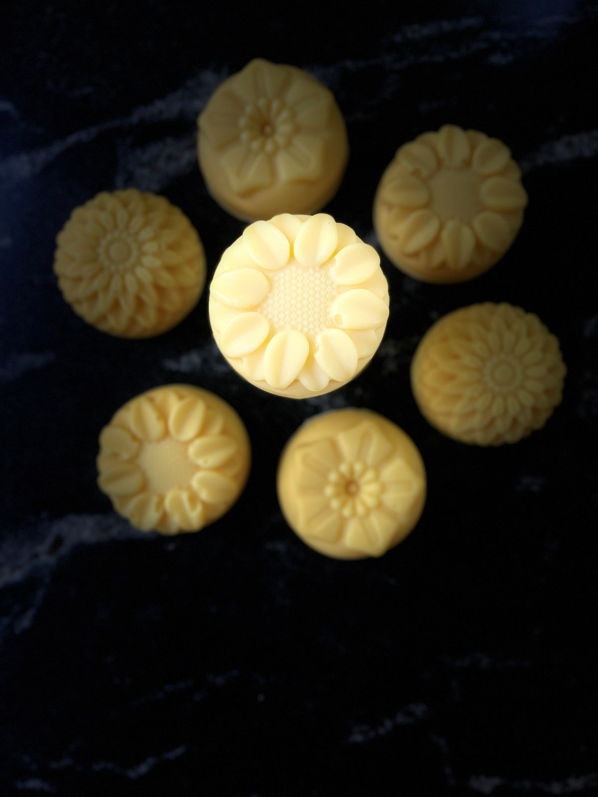 Seven pale yellow flower-shaped soap bars arranged on a dark marble-like surface, with one central soap bar highlighted by brighter lighting.