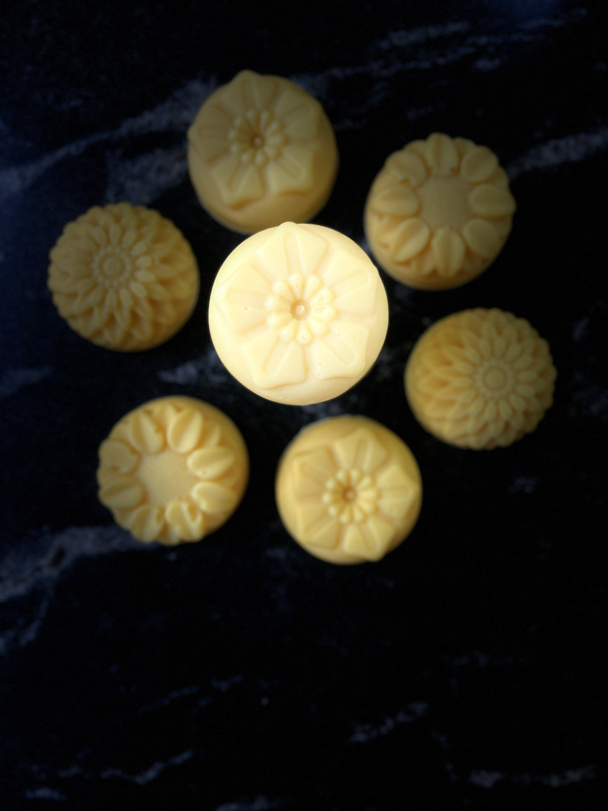 Seven pale yellow flower-shaped soap bars arranged on a dark marble-like surface, with one central soap bar highlighted by brighter lighting.