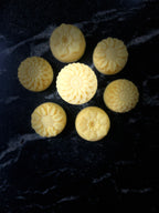 Seven pale yellow, flower-shaped wax melts arranged on a dark, textured black marble surface.
