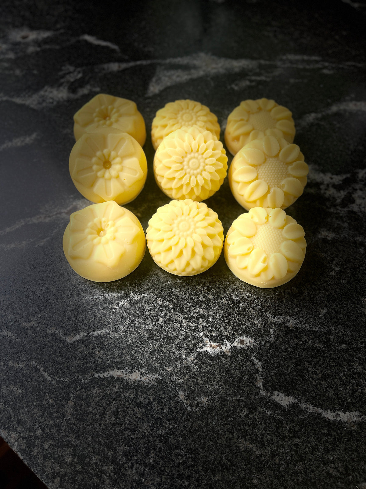 Nine pale yellow, flower-shaped handmade soaps arranged in a 3x3 grid on a dark, polished marble surface.