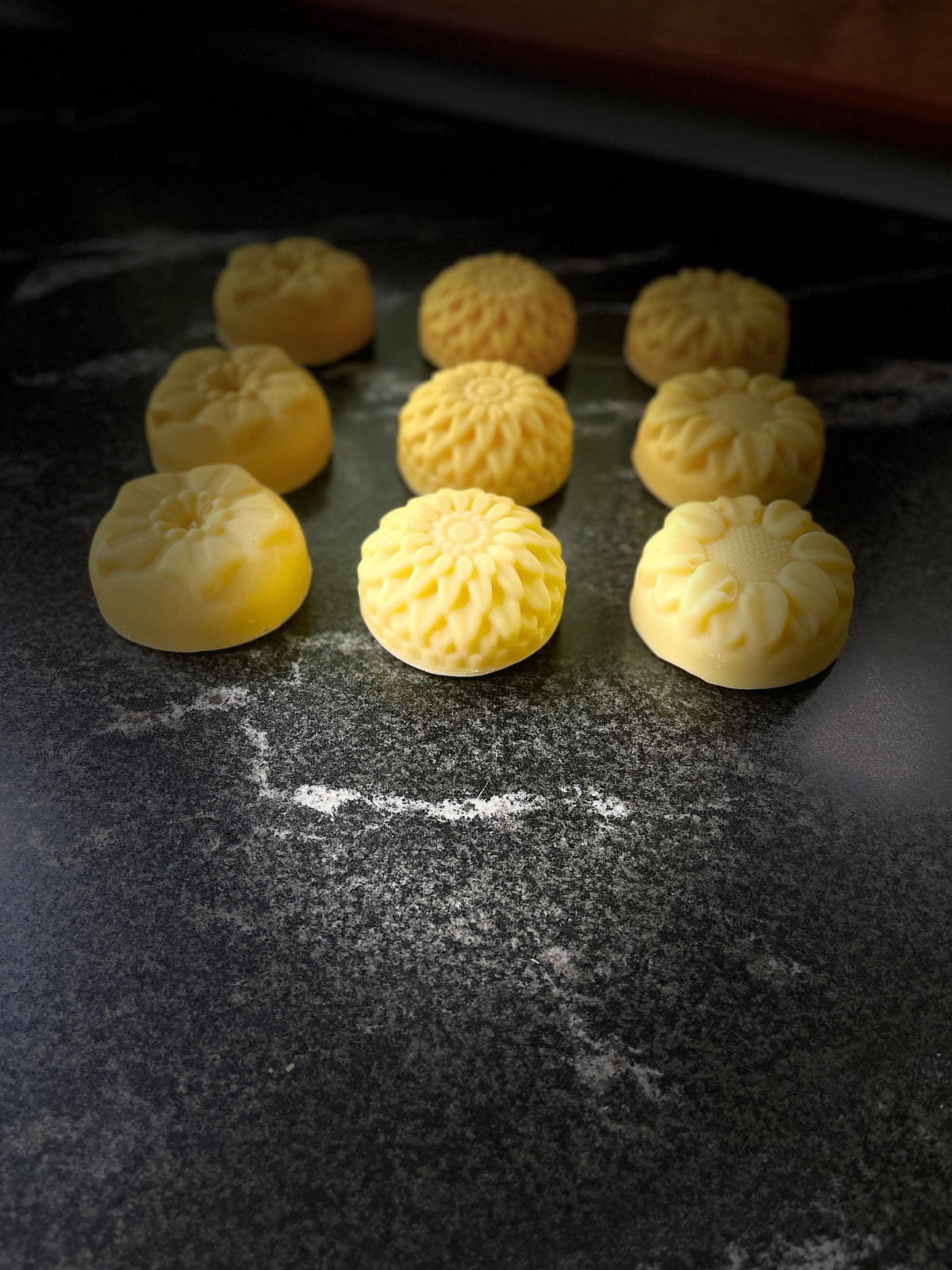 Nine round yellow homemade soaps shaped like flowers arranged on a dark surface.