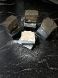 Four artisanal soap bars with a black layer and a taupe cream marbled patterns, displayed on a dark marble surface. There is a white silhouette of a tipped wolf in the bar. One bar in the foreground is stamped with the brand name 'Savi Suds'