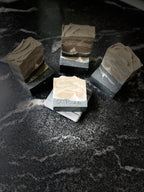 Four artisanal soap bars with a black layer and a taupe cream marbled patterns, displayed on a dark marble surface. There is a white silhouette of a tipped wolf in the bar. One bar in the foreground is stamped with the brand name 'Savi Suds'