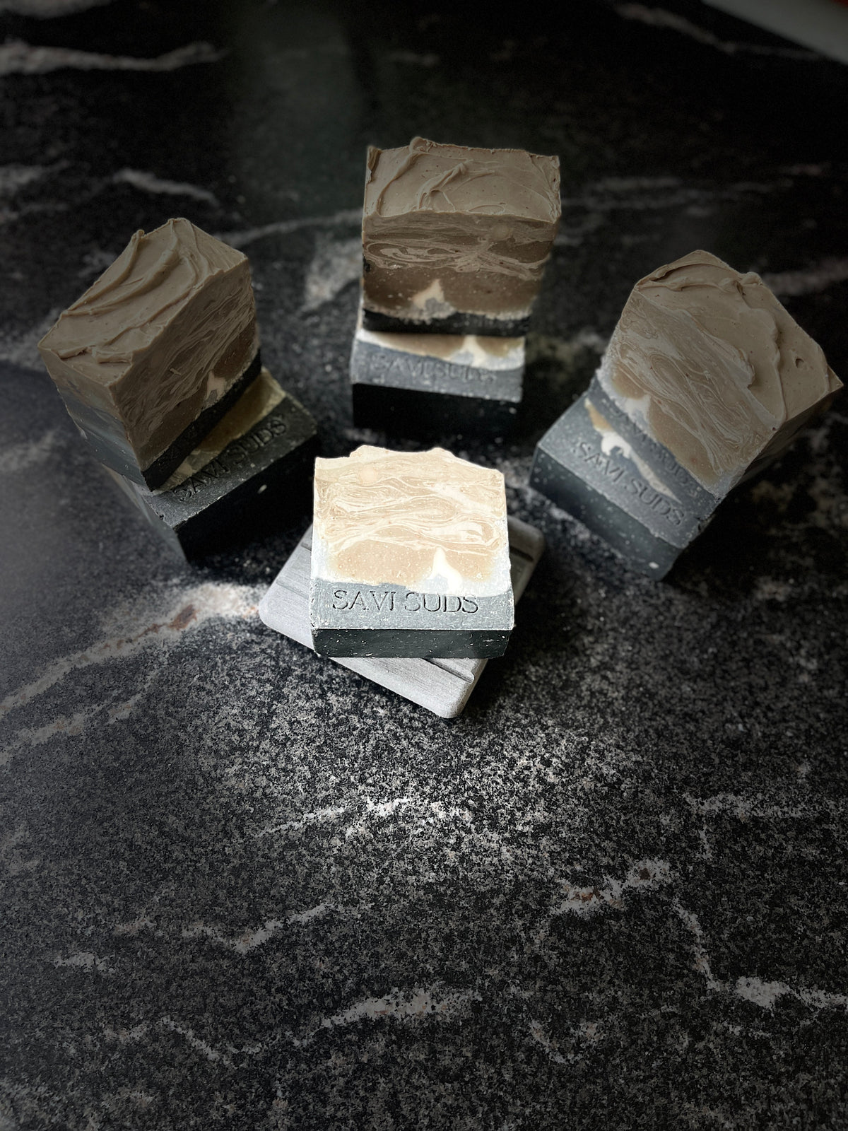 Four artisanal soap bars with a black layer and a taupe cream marbled patterns, displayed on a dark marble surface. There is a white silhouette of a tipped wolf in the bar. One bar in the foreground is stamped with the brand name 'Savi Suds'