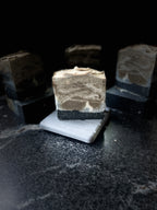 A styled product shot of artisanal layered soap bars. A central bar is displayed upright on a small grey stone, showcasing a marbled pattern of light brown, cream, and dark charcoal layers. A white silhouette of a tilted wolf is howling at the moon