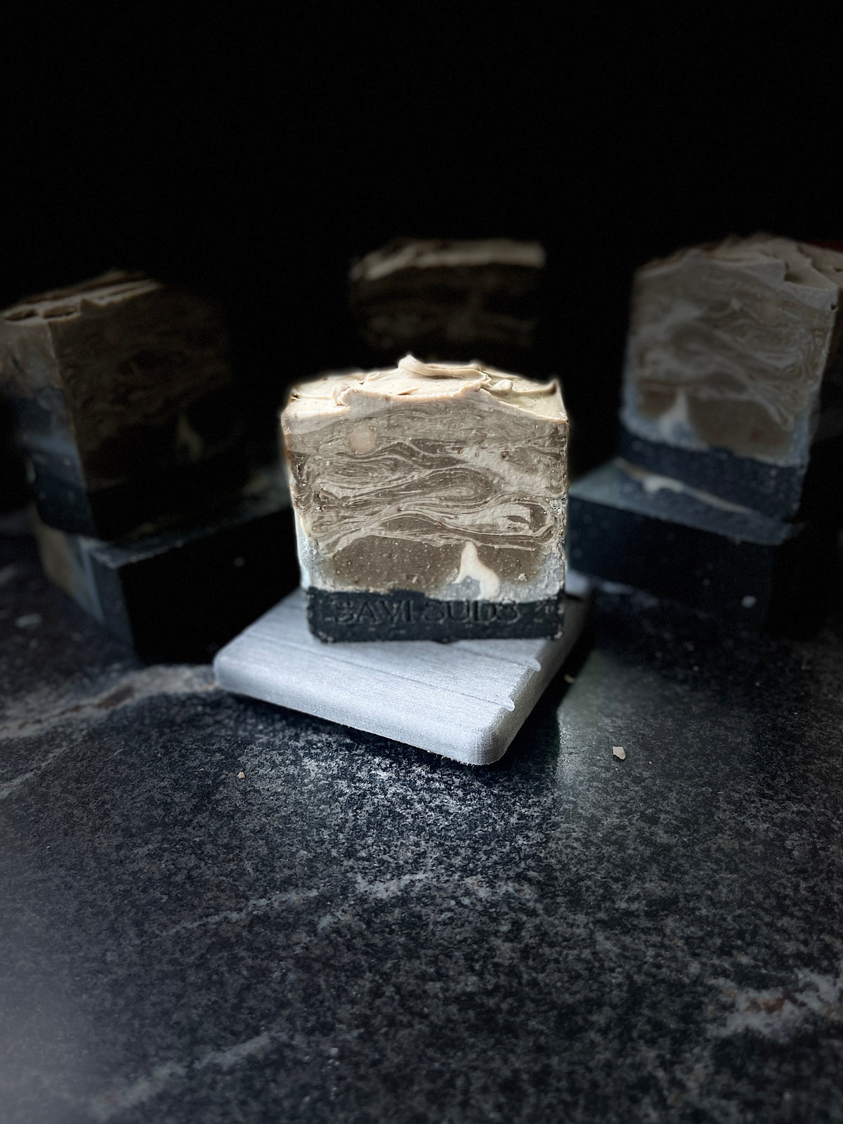 A styled product shot of artisanal layered soap bars. A central bar is displayed upright on a small grey stone, showcasing a marbled pattern of light brown, cream, and dark charcoal layers. A white silhouette of a tilted wolf is howling at the moon