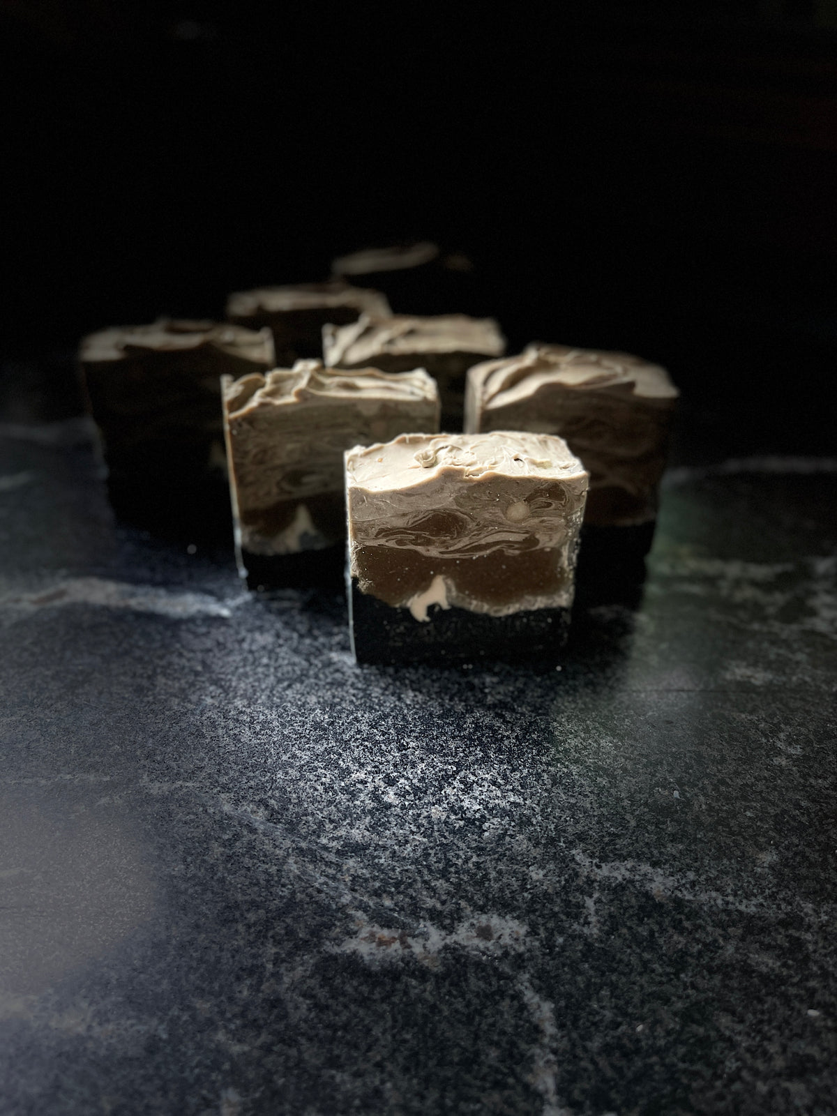 Four layered soap bars with dark brown and light beige swirls, arranged on a dark textured surface against a black background.