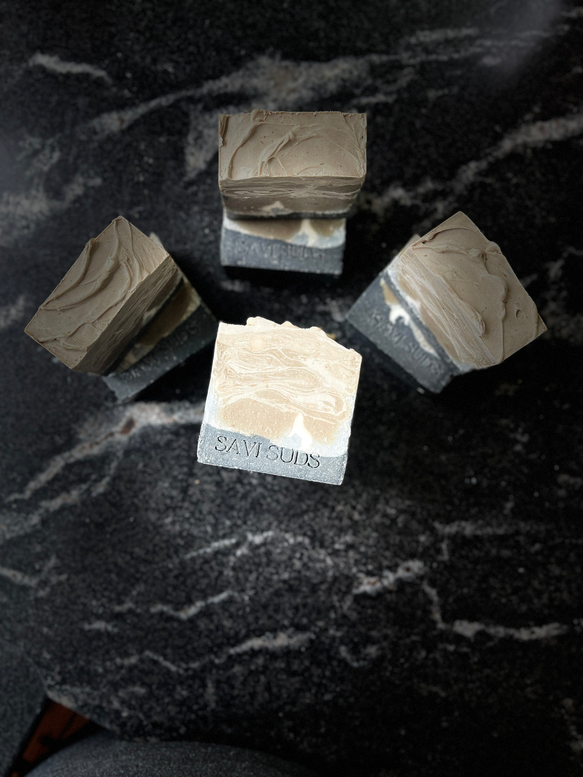 Four artisanal soap bars with textured taupe and cream marbled tops and dark gray stone bases, arranged on a dark marble surface. One bar in the foreground is engraved with the brand name 'SAVI SUDS'.